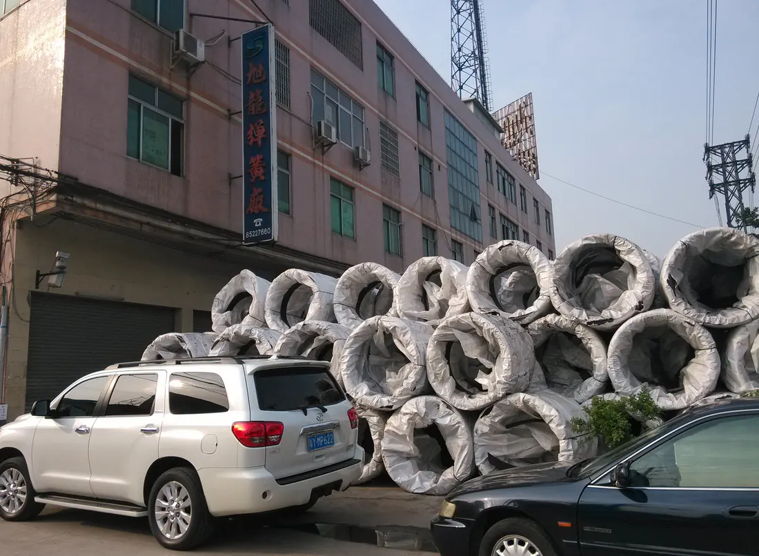 整卷日本神户制钢弹簧钢丝到厂卸车，卷料外覆白色防护袋，堆放在旭龙弹簧厂区门口，背景可见旭龙弹簧厂招牌